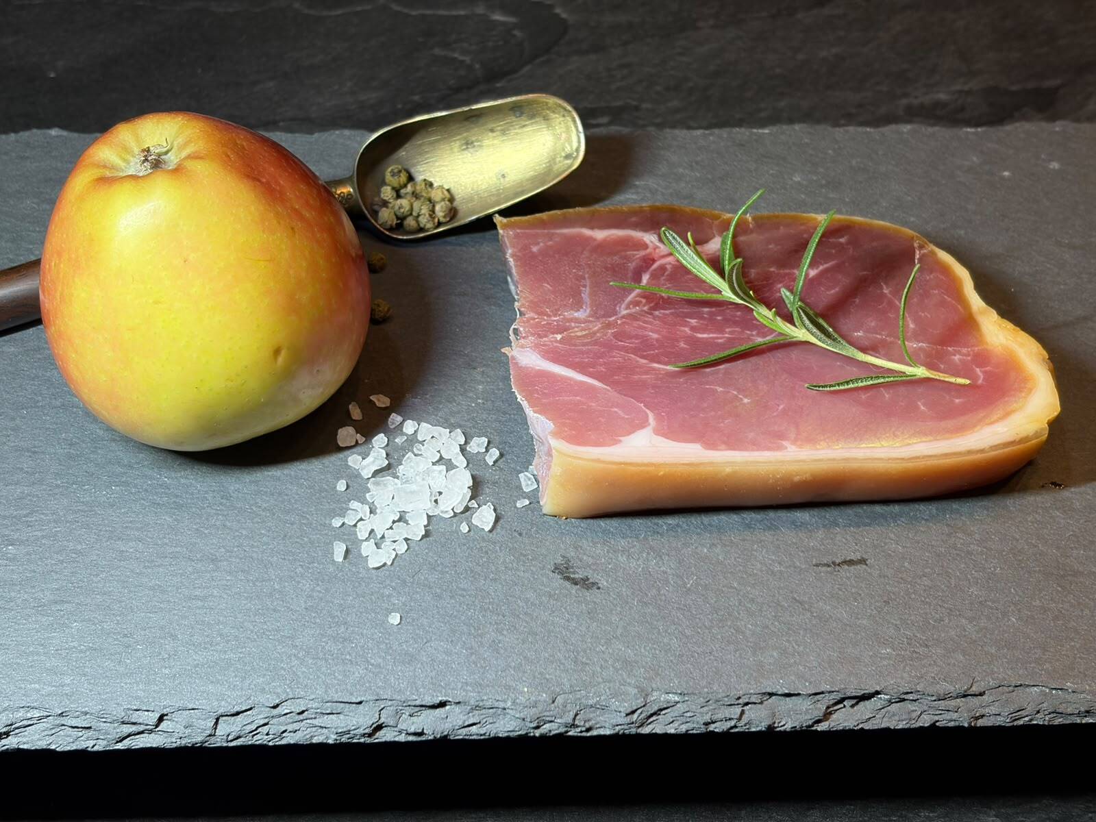 An apple, prosciutto, rosemary, green peppercorns, and sea salt on a slate board