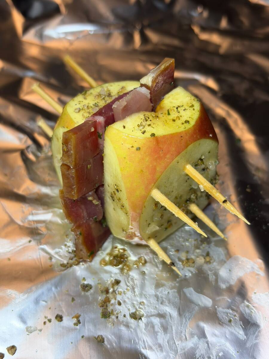 An apple filled with prosciutto and pinned with skewers, sitting on aluminum foil