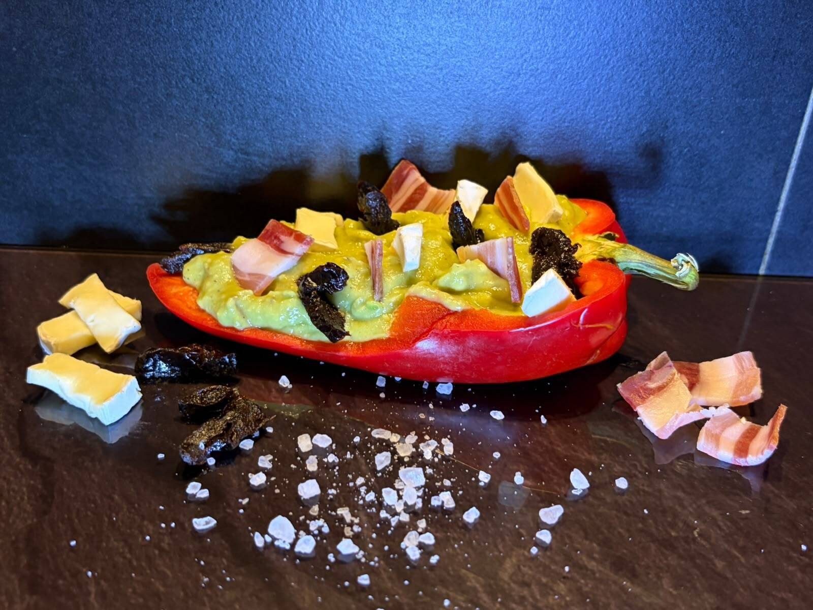 A halved red pepper filled with avocado spread and topped with pancetta, dried plum, and brie
