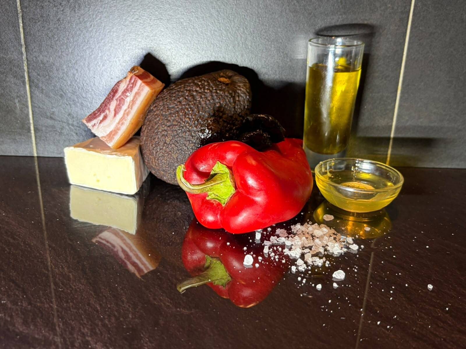 Brie, pancetta, an avocado, a red pepper, olive oil, and sea salt arranged on a dark surface