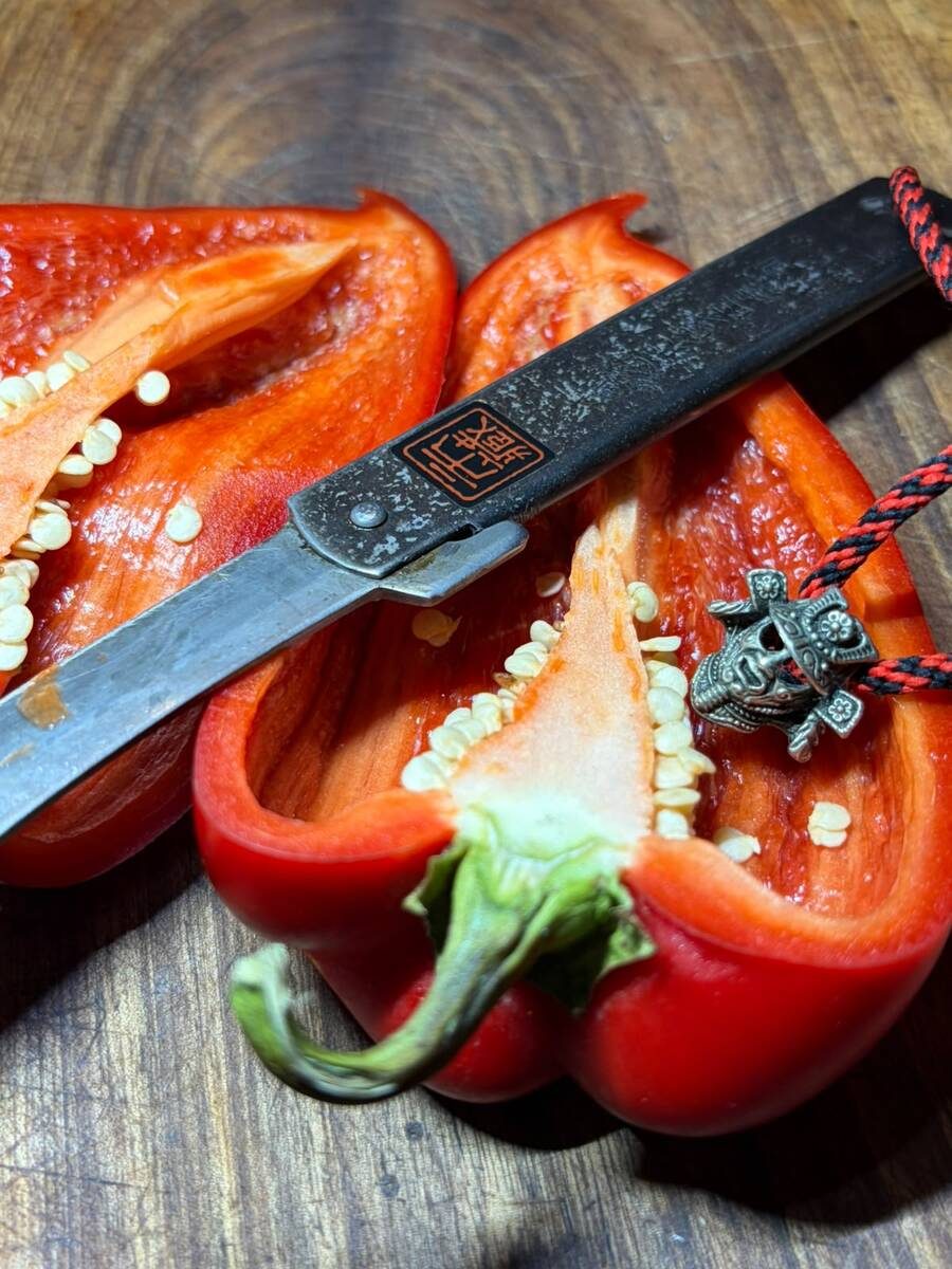 An Ichizo Honpo Higonokami, with its red cord and samurai charm, resting on a red pepper split lengthwise with its seeds showing